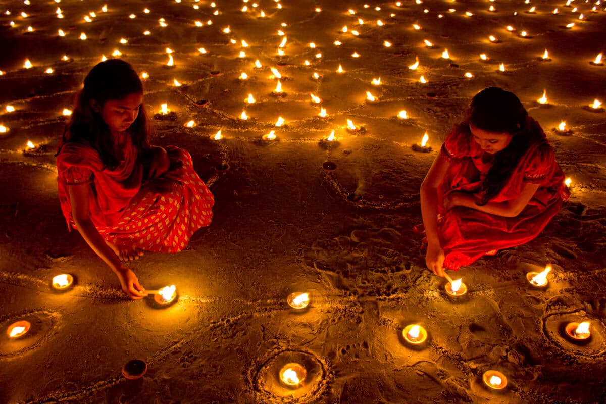 Two girls lighting rows of clay diyas at dusk